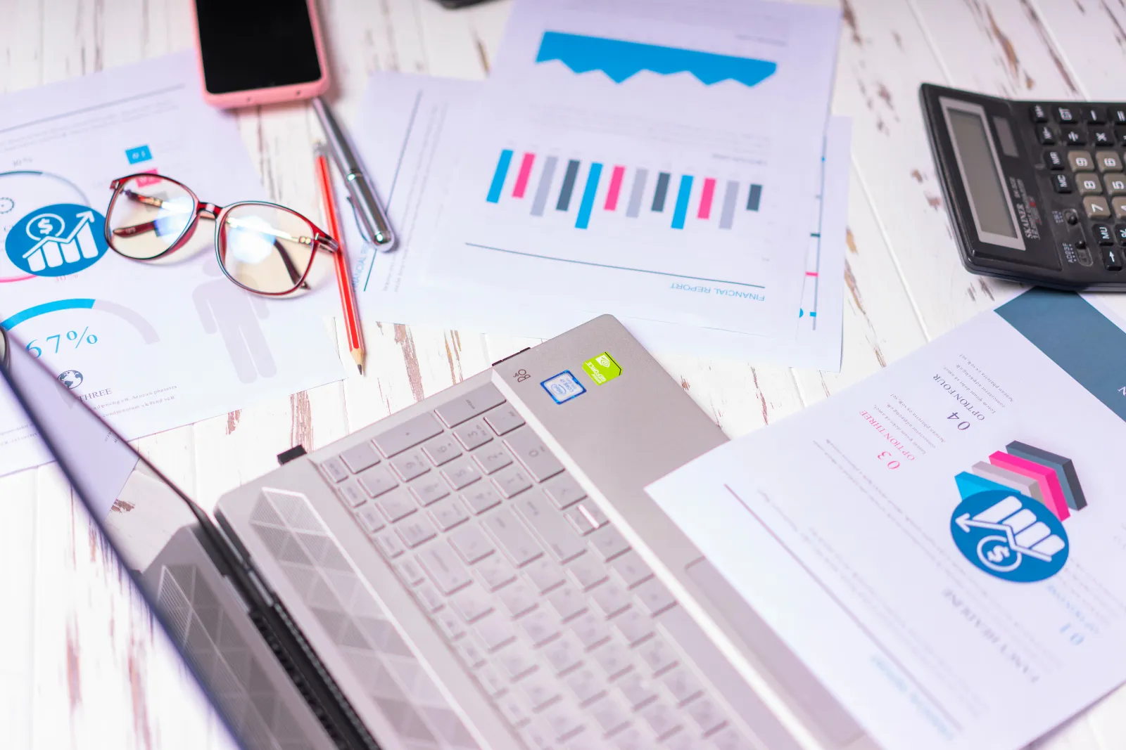 Printed financial reports, a calculator and a laptop spread across a desk