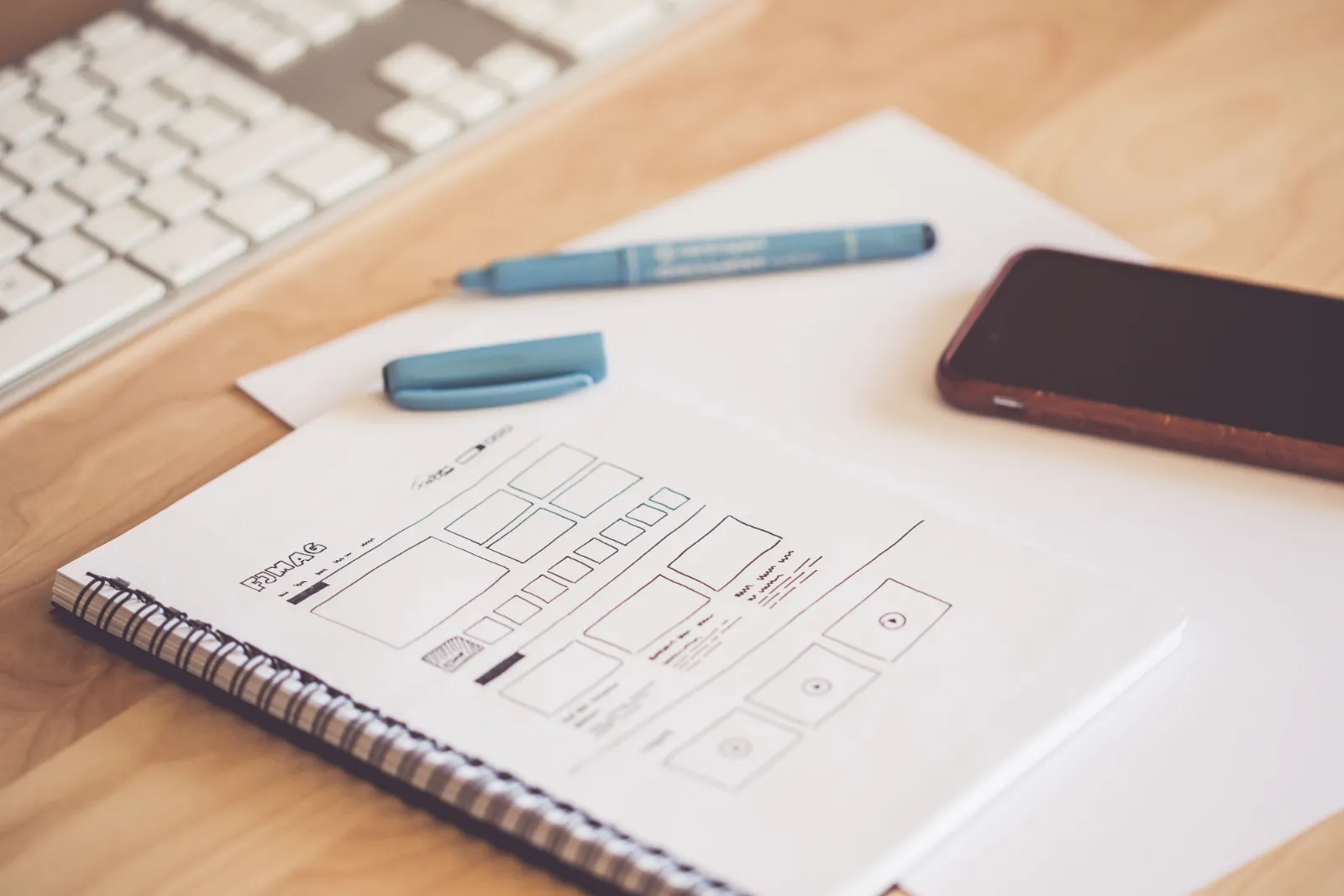 A hand-drawn app wireframe in a spiral notebook beside a pen, keyboard and phone