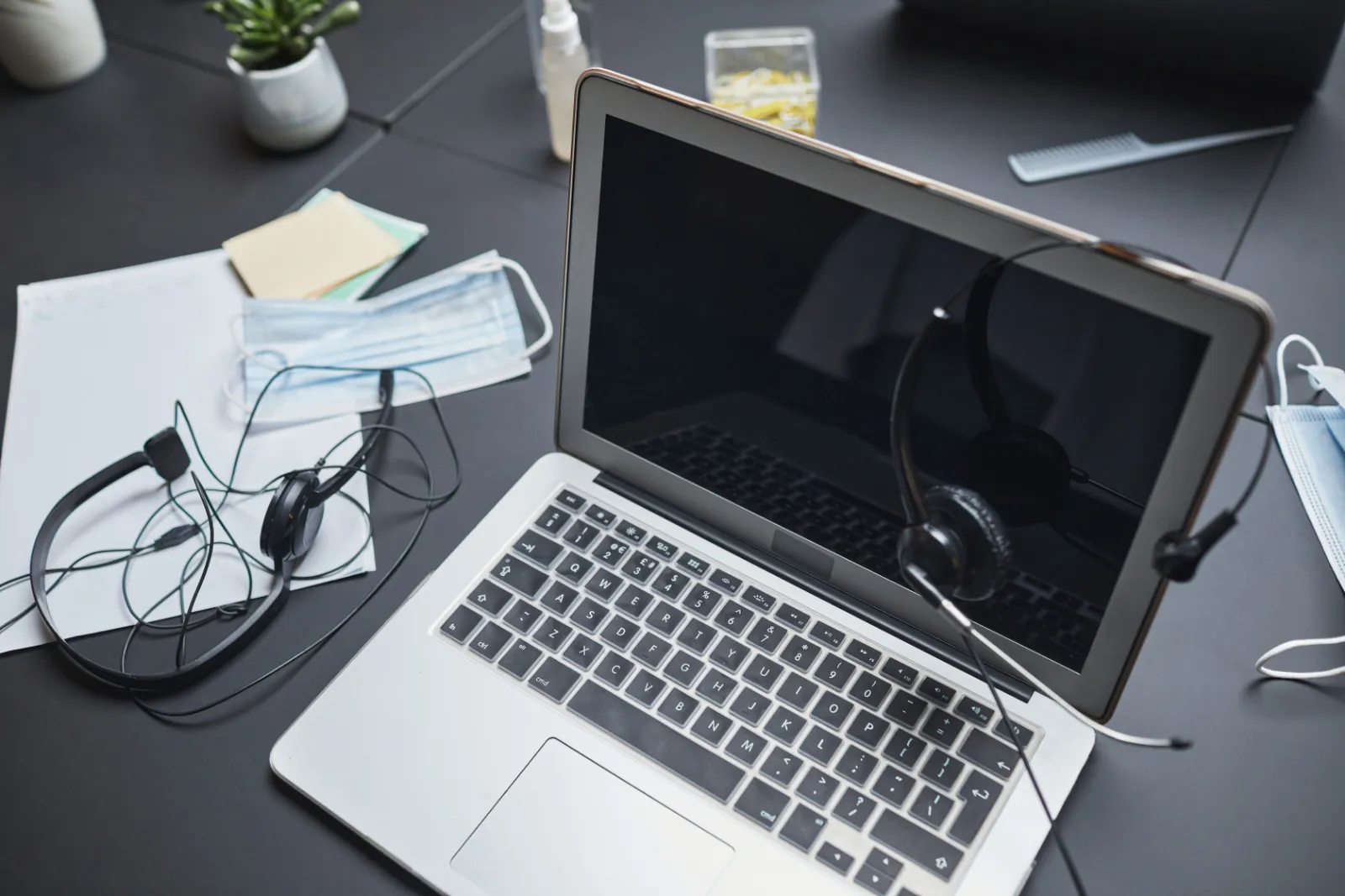 An open laptop on a dark desk with a calling headset, notes and sticky notes beside it