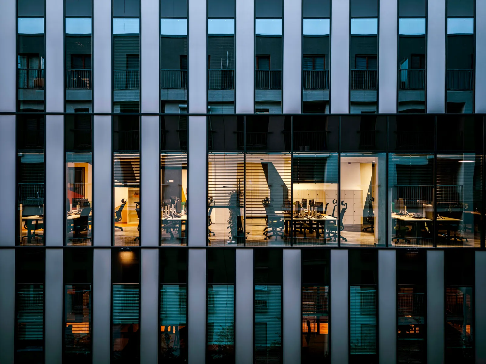 A modern office building at dusk, with one floor of desks and monitors lit behind the glass