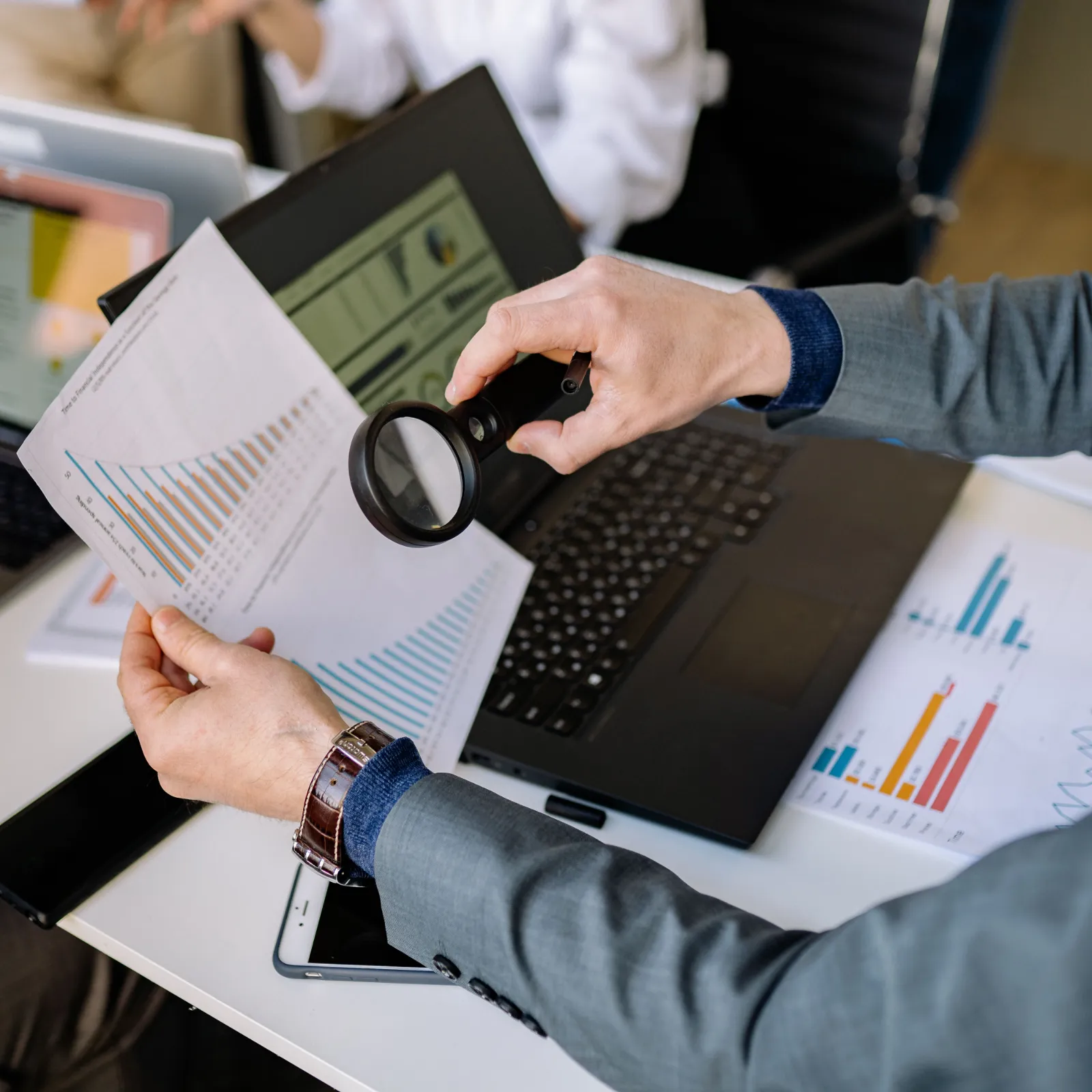 A hand holding a magnifying glass over printed charts beside a laptop