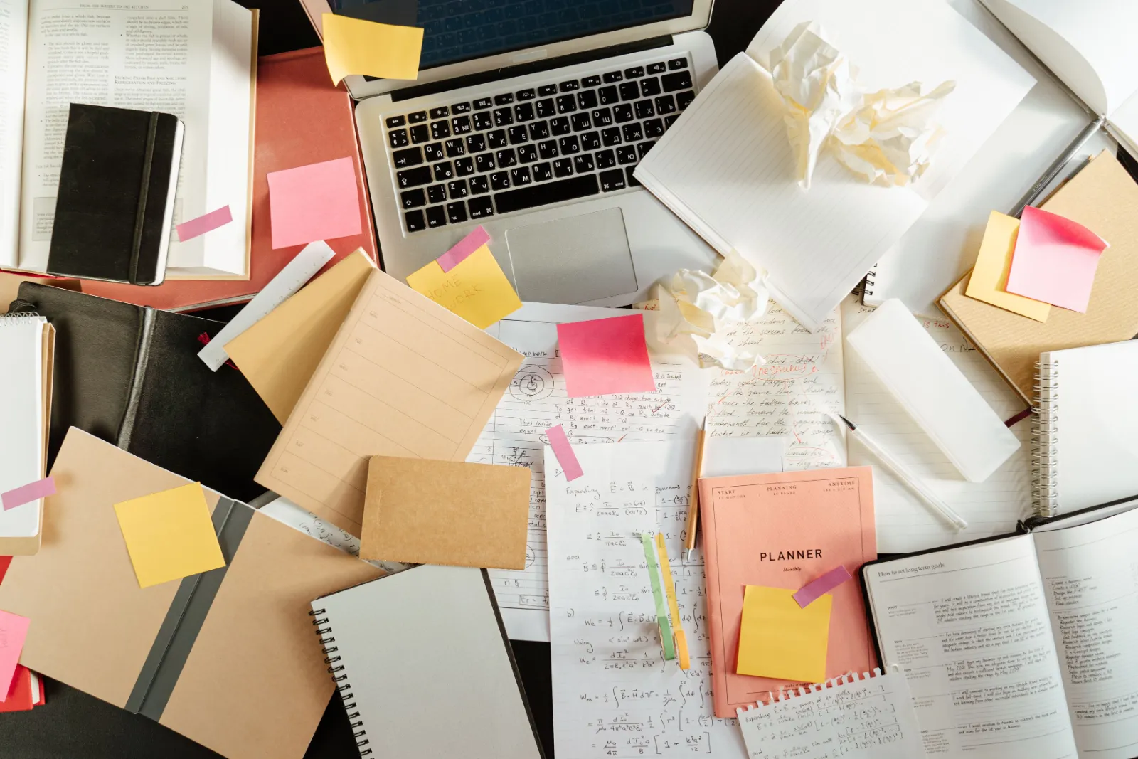 A cluttered desk covered in notebooks, sticky notes and crumpled paper around a laptop